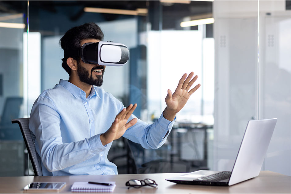 A man in a VR headset raising his hands at a desk with a laptop.